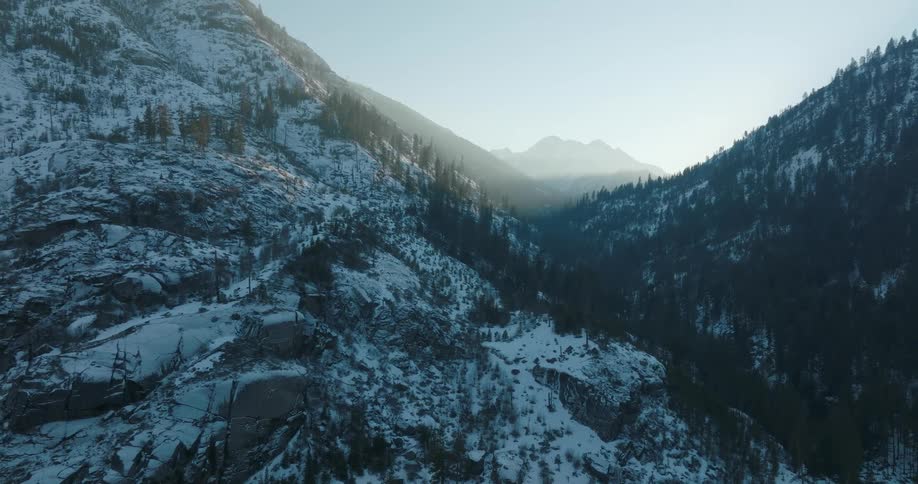 Pullback tilt up reveal from the boulders and rock face of a snow covered mountain slope and deep forested valley below