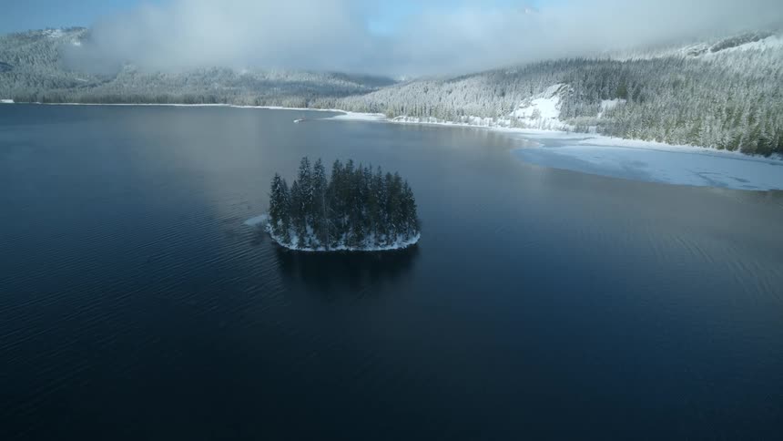 A small island covered with snow dusted fir trees sits in the middle of a cold mountain lake