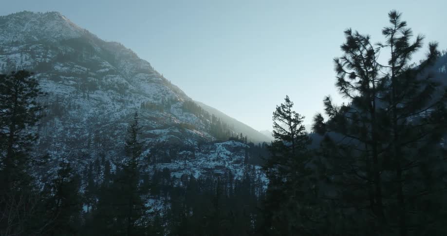 The silhouette of evergreen trees cross frame in front of a beautiful snow dusted mountain valley in the distance