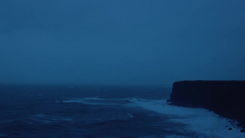 Waves flow across ocean to crash against cliff on foggy day