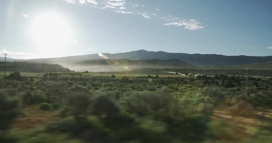 The sun glowing brightly in the sky above grasslands with trees and brush and a mountain on the horizon