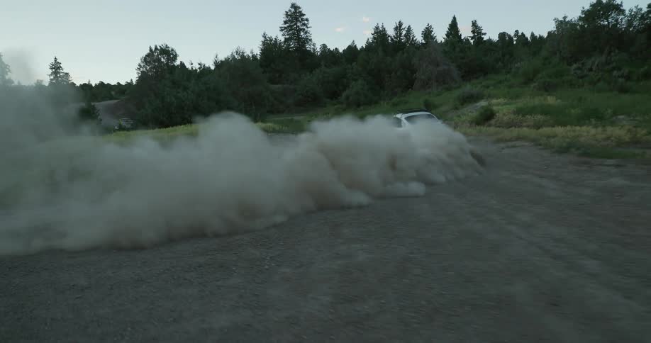 A man quickly drives a car around a corner on a dirt road in a rural area and dust flies behind as he turns