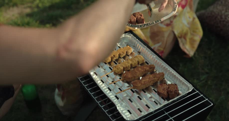 A woman holds a tray with food and a man takes the food and puts it on a grill to cook