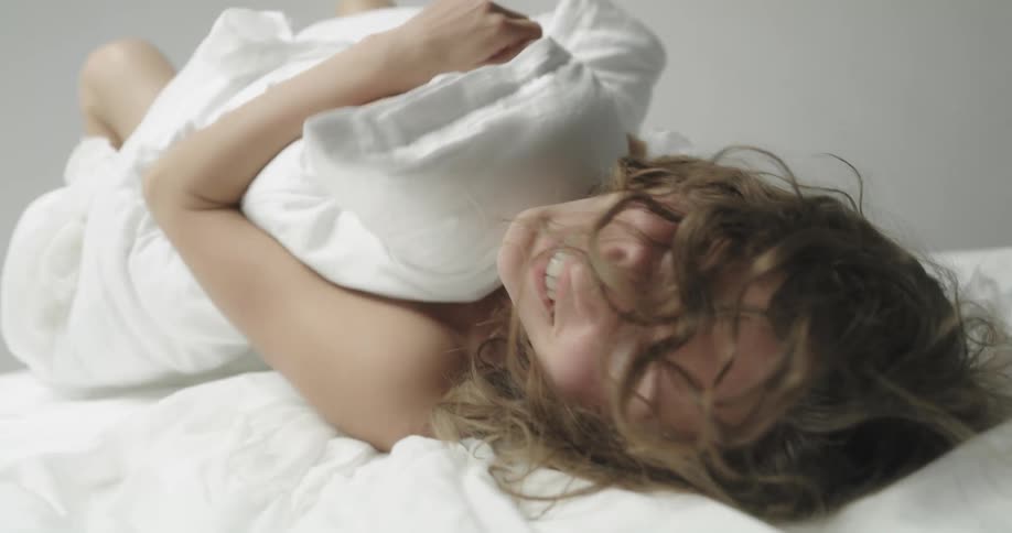 A naked woman smiles and laughs as she rolls around in bed sheets