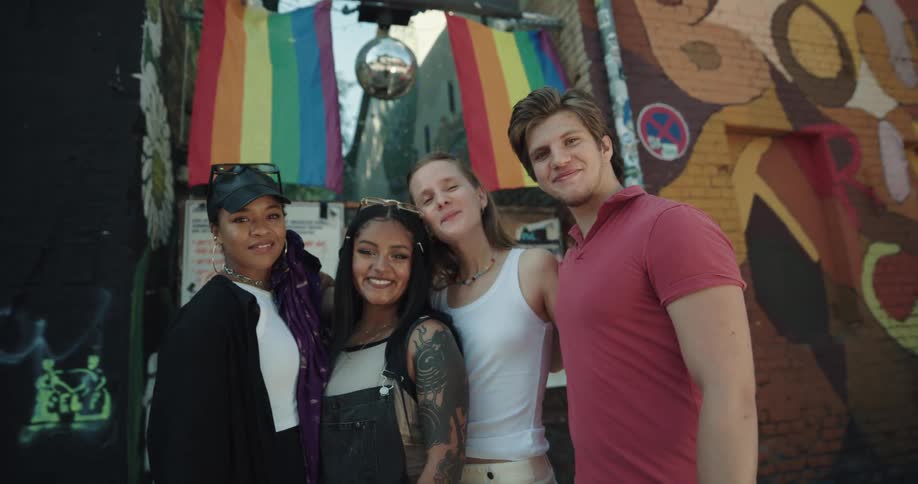 A group of friends smile as they pose in front of rainbow flags hanging between downtown buildings