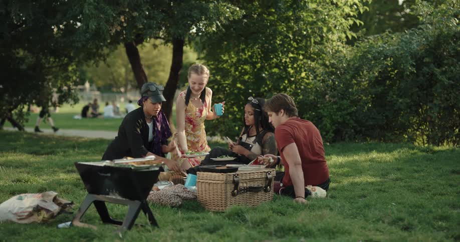 Friends grill and have picnic in park as musician walks past in background carrying an instrument case