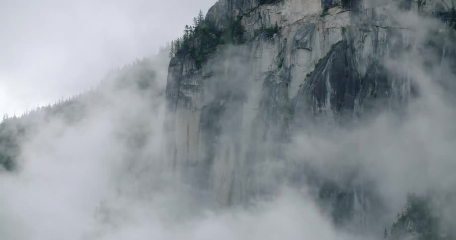 Mist rises up the side of a sheer rock cliff face and across a hill on a cloudy day