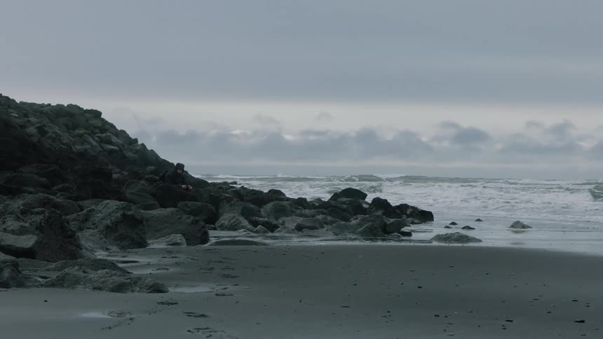 Person wearing coat sits among rocks on shore as waves roll onto beach