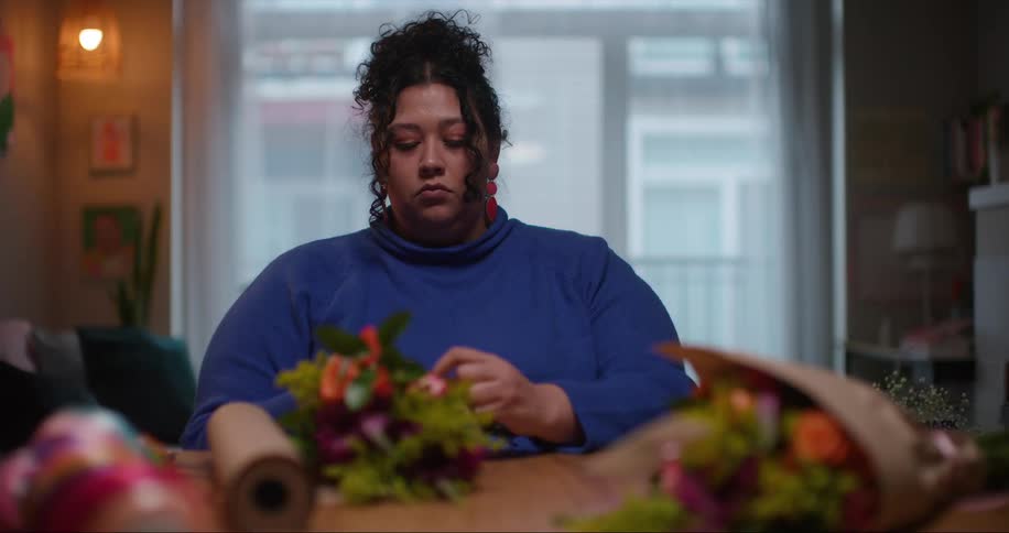 A woman sitting at a table in her apartment tying ribbon around flower bouquets