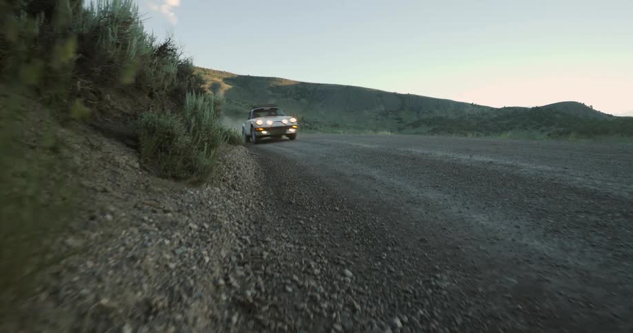 An automobile travels around a curve on a dirt road and there are mountains in the distance