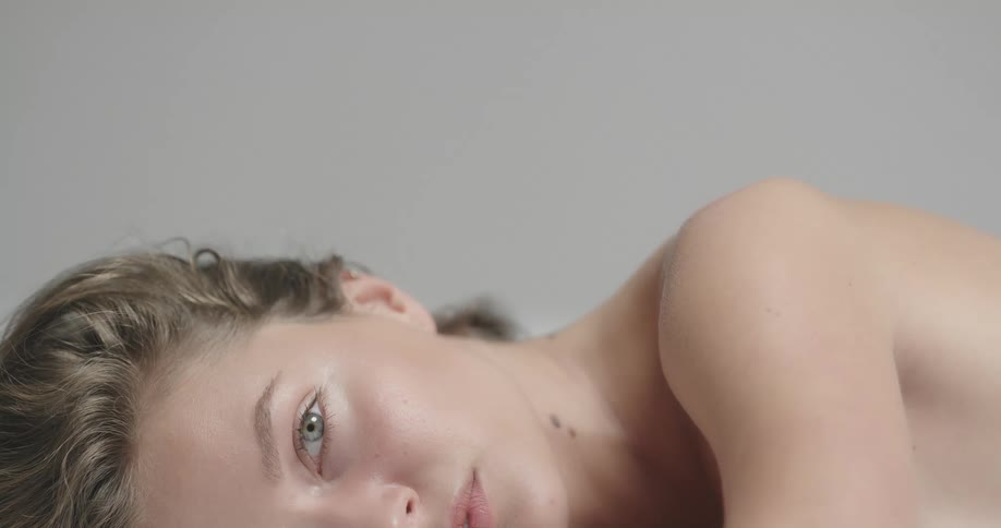 Topless female model with brown wavy hair lies in prone position looking at camera