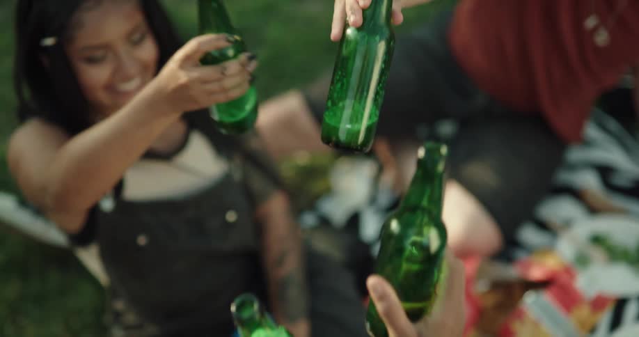 A group of friends hold up their beer bottles for a toast while picnicking in a park