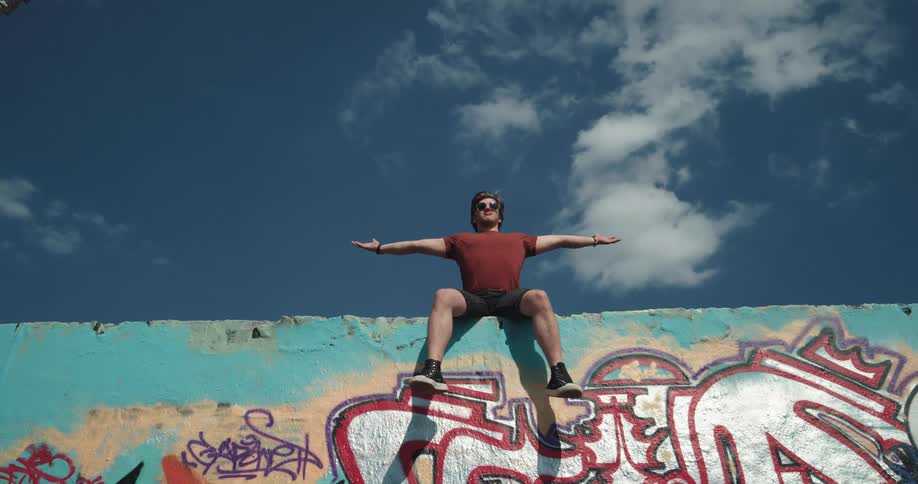 A man sitting on the edge of a wall swinging his legs stretches his arms out as if he is king of the world