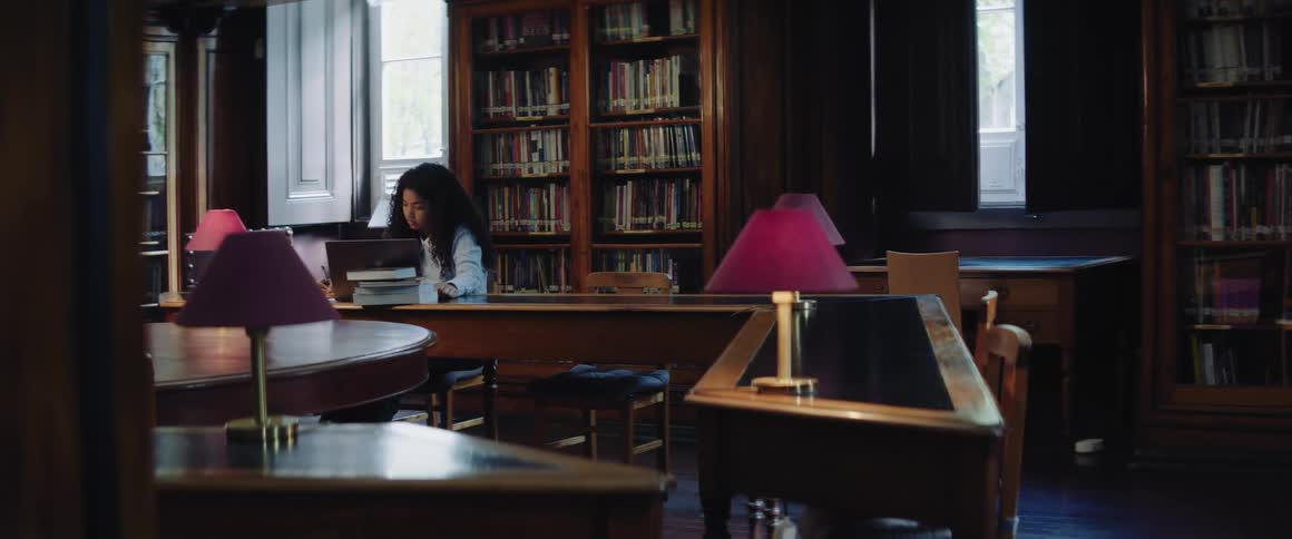 A teenage girl is sitting at a table in a library behind a stack of books and a laptop computer