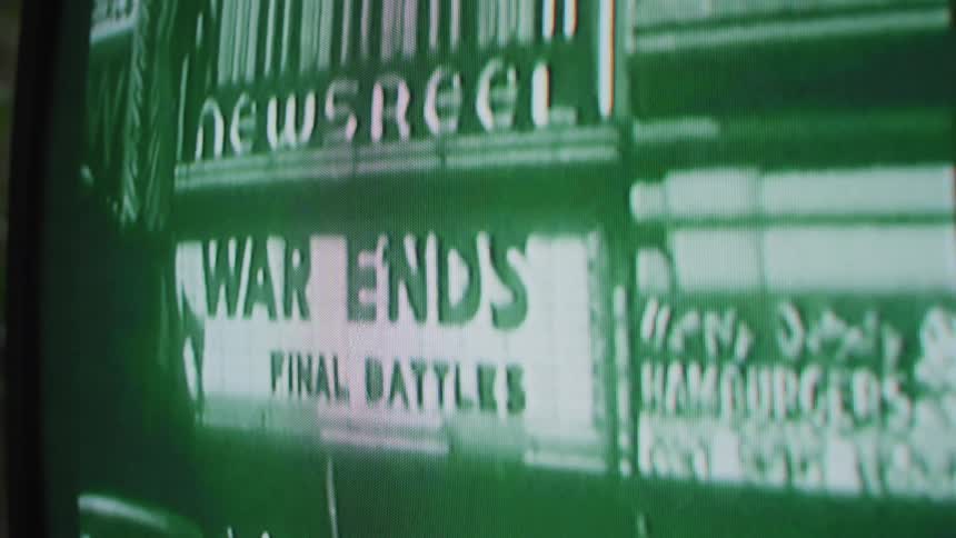 A television screen displays an archival scene of a city theater marquee advertising newsreels and war footage