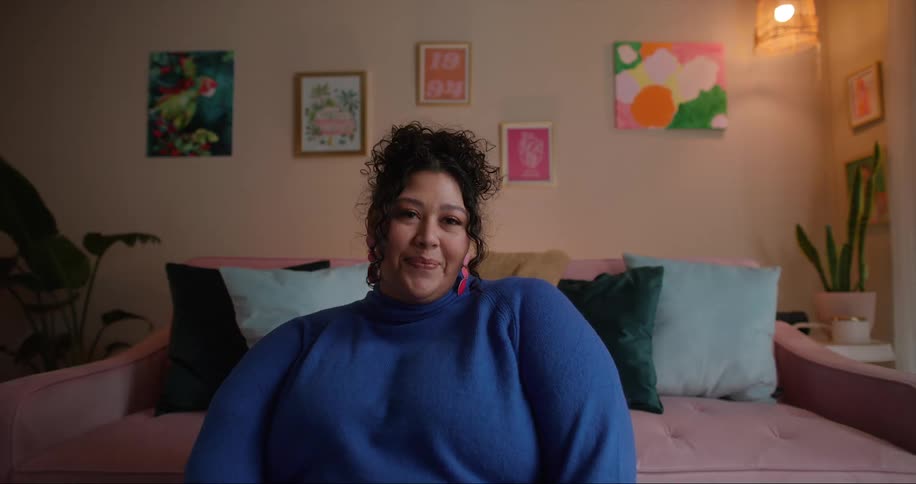Obese woman with curly hair and blue sweater sits by pink couch smiling at camera