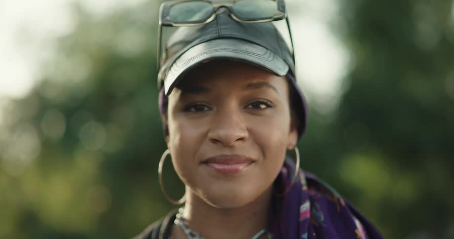 Woman with sunglasses black cap hoop earrings purple scarf and necklace smiles at camera