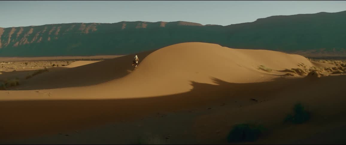 The sun shines brightly upon a man running up a hill in the desert and there are mountains in the distance