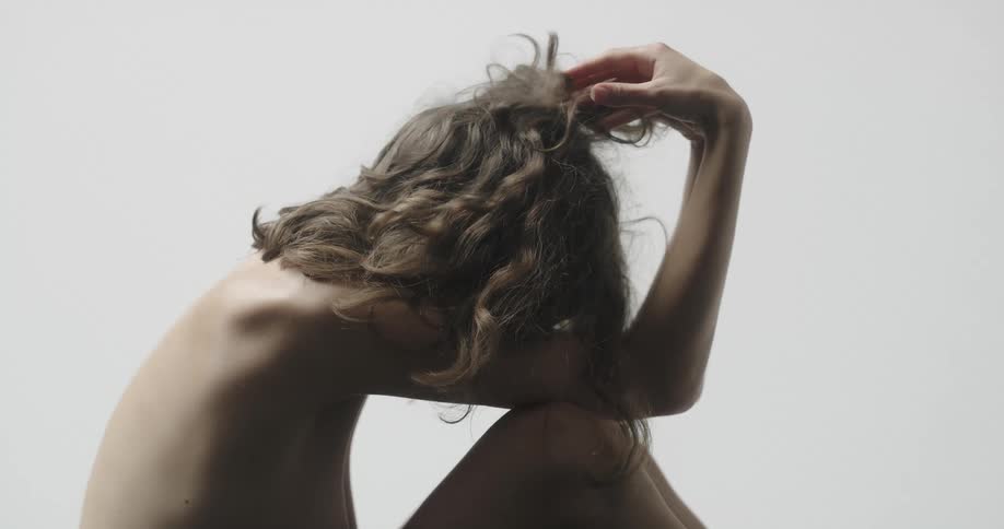 Topless female model sits on the ground touching her brown wavy hair
