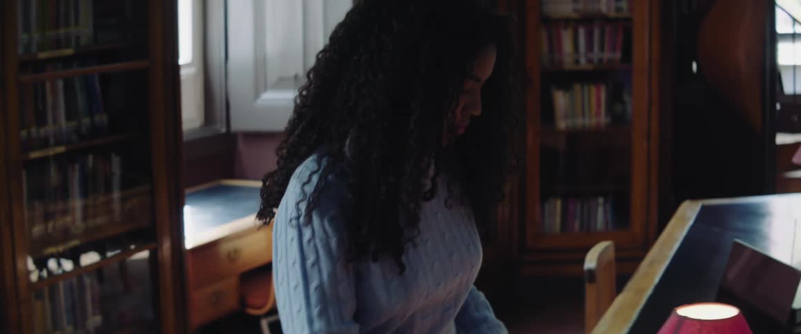 Female college student takes book from bookcase and sits down to look through it at table with open laptop