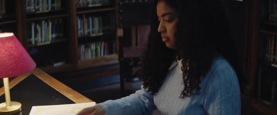 Woman with curly hair and turqoise sweater sits at desk holding book in the library