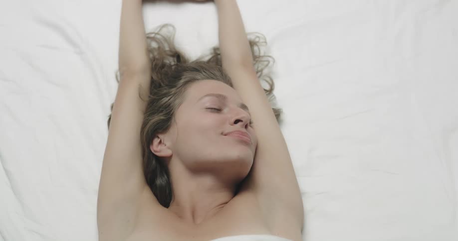 Topless female model with brown wavy hair lies down and stretches her arms