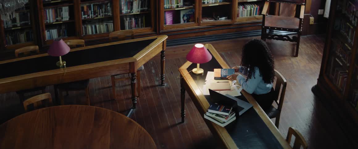 A young woman takes a book off of a shelf and sits down to do school work