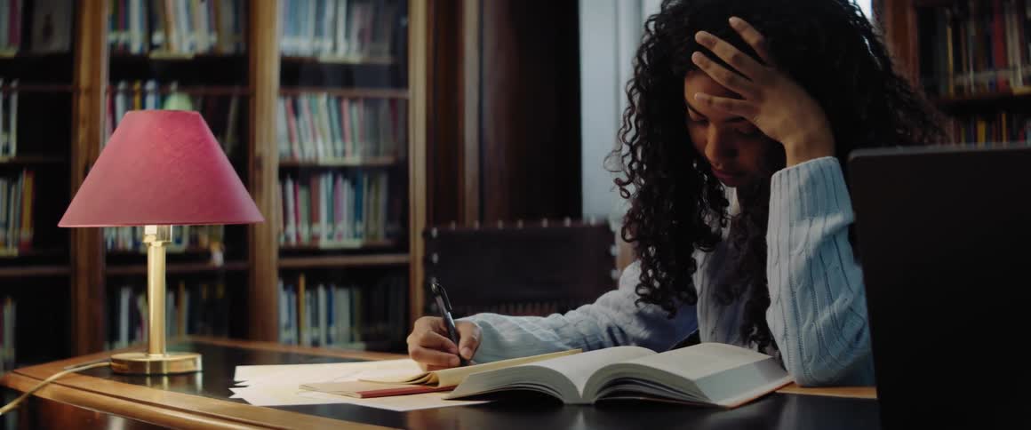 A teenage girl is sitting at a table in a library taking notes from an open book in front of her