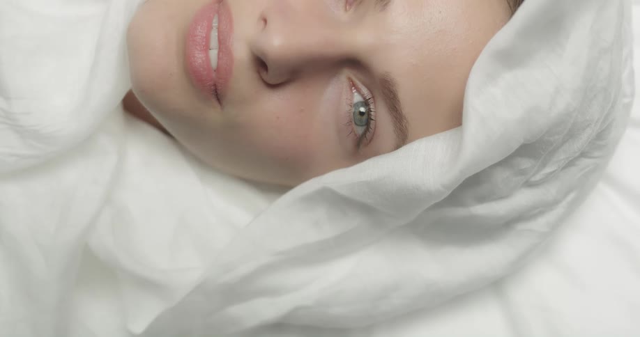 A woman smiles while lying in bed with a sheet wrapped over her head