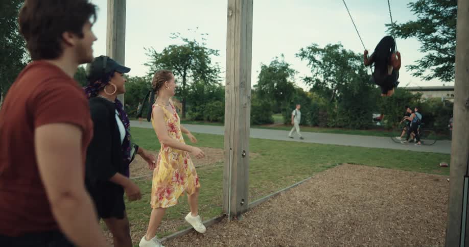 A teenage boy pushes a girl on a swing set at a busy park as her friends laugh and others walk by