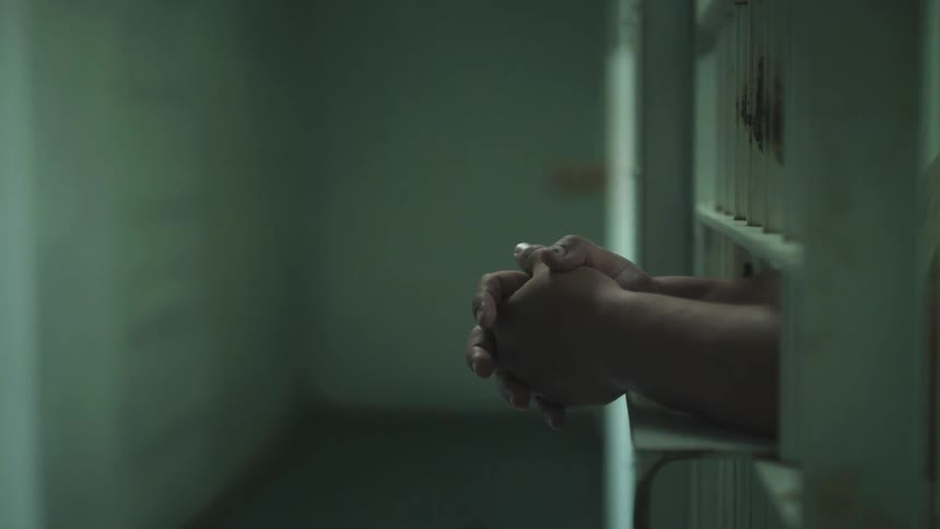 Close up of a mans hands hanging out between bars in a prison cell