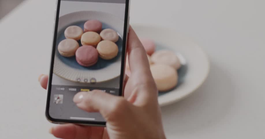 Cookies are on a plate sitting on a table and a woman takes a picture of them with her phone