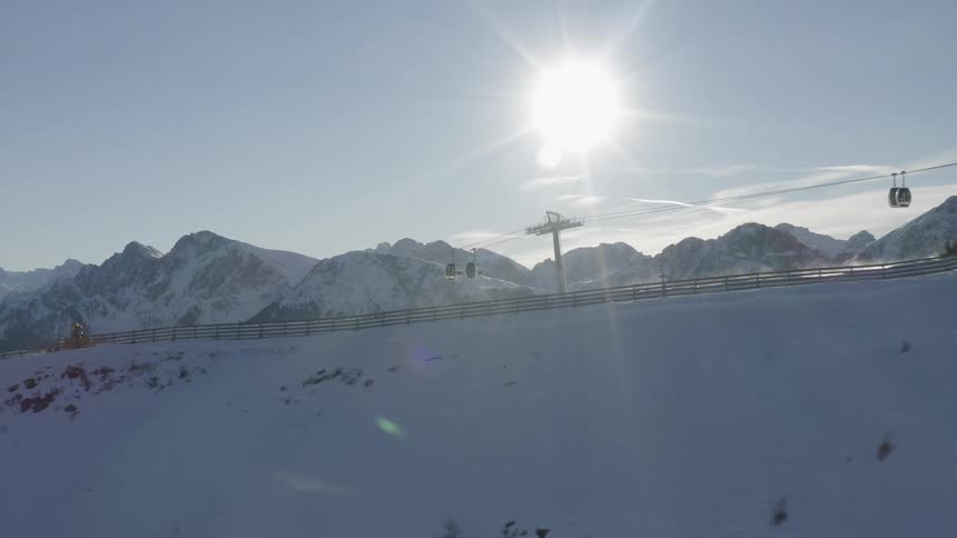 The sun is shining brightly as gondola lift cars travel over the top of a snow covered hill as someone skis and there are mountains in the distance