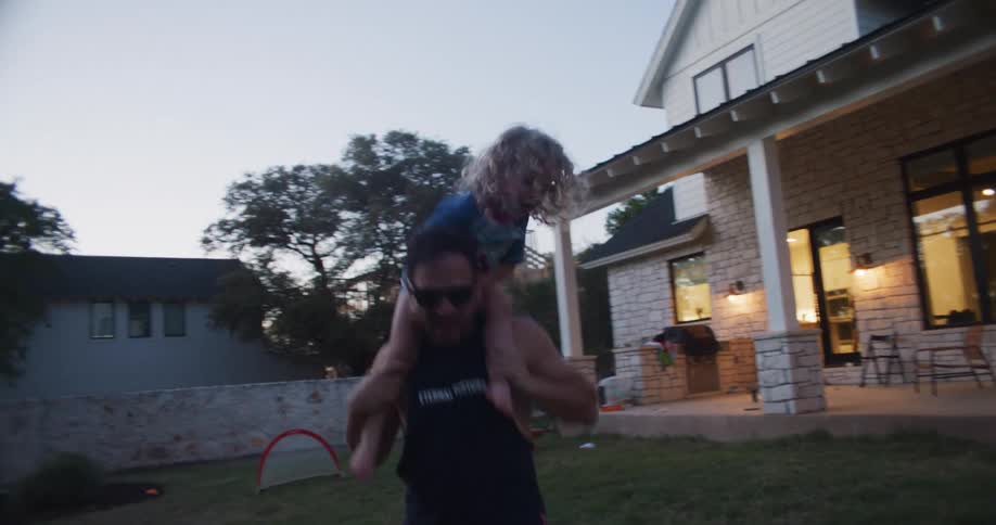 Father carries boy on his shoulder while mother cooks in kitchen