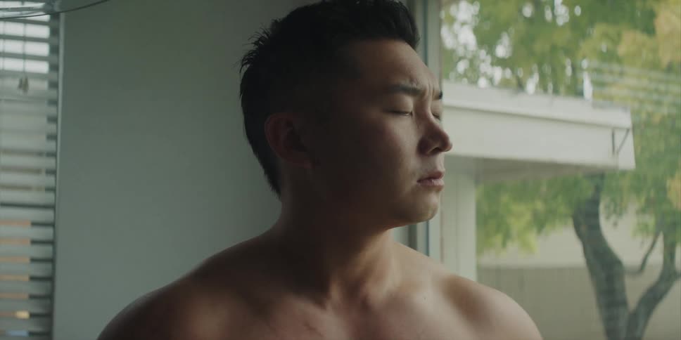 A bare chested asian man is standing near a window drinking a cup of coffee