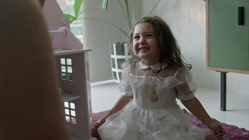 A little girl wearing a white dress is sitting on the floor laughing with her grandmother