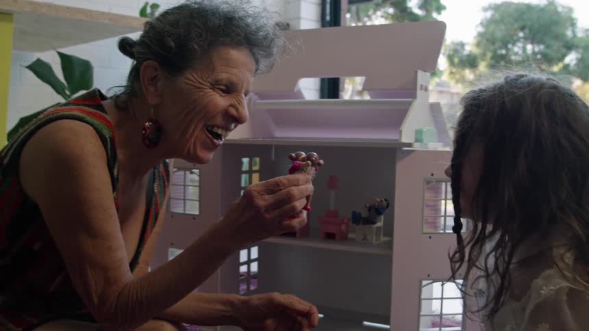A little girl wearing a party dress laughs as she plays with her dollhouse with grandma