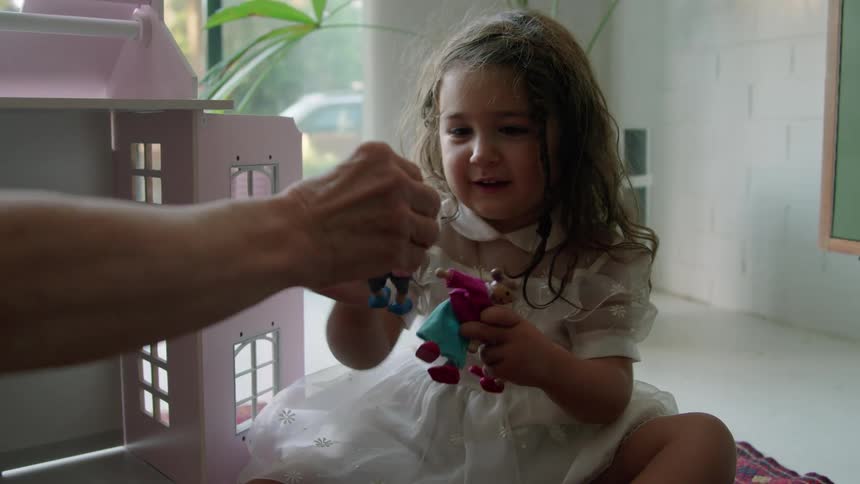 A little girl wearing a white dress is sitting on the floor and an adult is handing her dolls