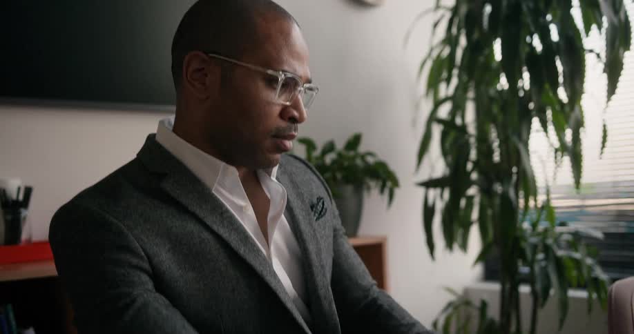 A man removes his eyeglasses from a case and puts them on his face before beginning to type on a laptop as he sits at a table in an office during the day
