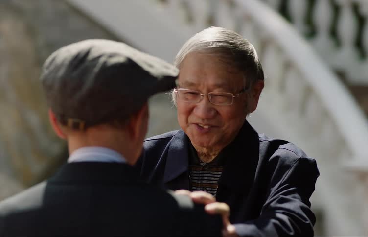Two older asian men are outside talking and one man puts his hand on the other mans shoulder