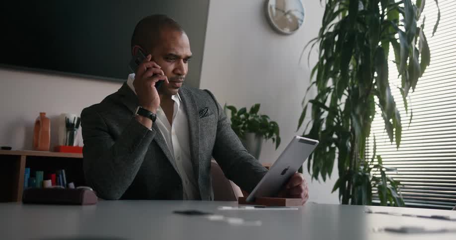 Man sits in office viewing ipad while he answers cellphone