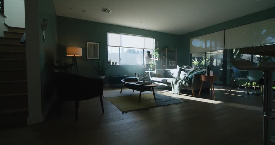 A woman lifts the shade on her large window bringing the bright sunshine into her living room