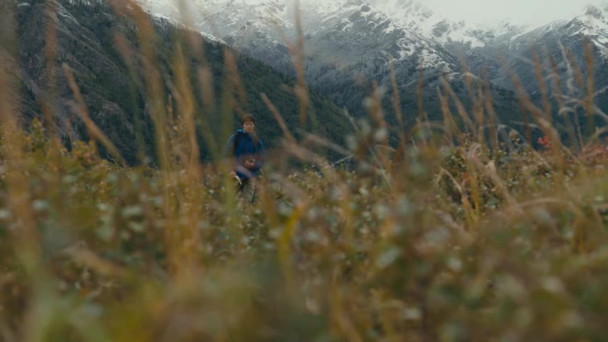 Photographer hikes across meadow with forested and snow covered mountains in the background