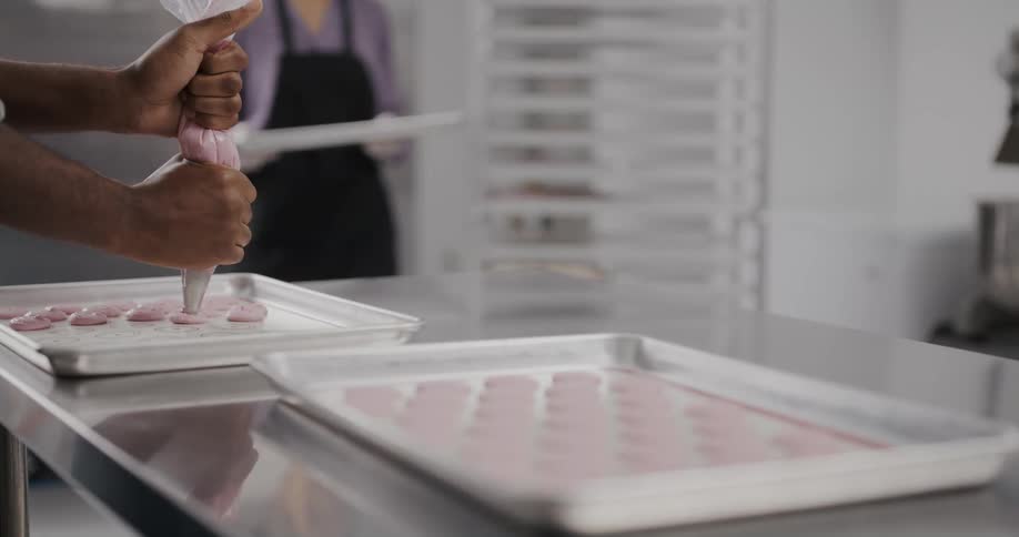 A woman removes a tray from a cooling shelf and places them on the counter where a baker is piping more onto trays