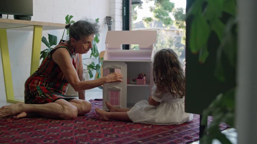 Old woman with striped dress plays with toddler girl with wavy hair and white dress next to pink model house sitting on red carpet in living room at home