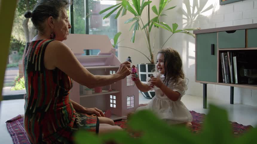 An elderly woman plays with a dollhouse with a young girl as they sit together on the floor indoors during the day