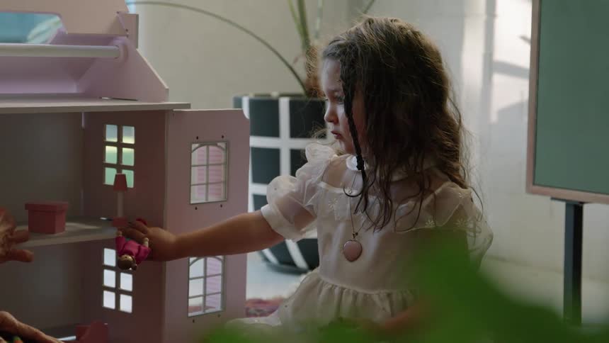 A little girl smiles as her mom puts a doll in a dollhouse
