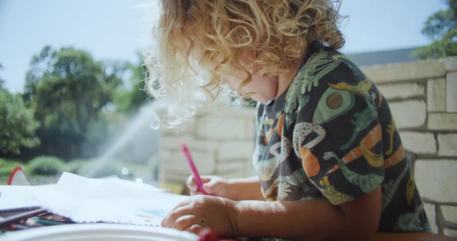 A little boy is coloring paper with a crayon as he sits on a balcony