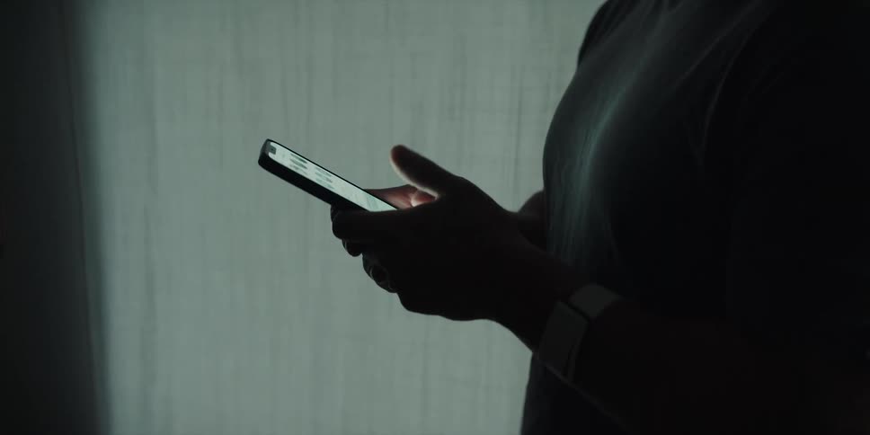 Man with shirt using smartphone is silhouetted next to curtains at dusk