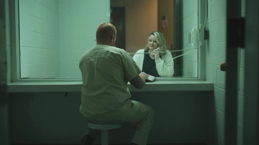 A woman is talking on a phone to an inmate she is visiting and sees though the glass
